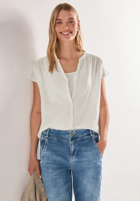 Vrouw met witte blouse en spijkerbroek, staand voor een lichte muur met handen in haar zakken.
