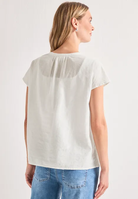 Vrouw van achteren in een wit kort mouwtoppje en een blauwe jeans voor een lichte achtergrond.