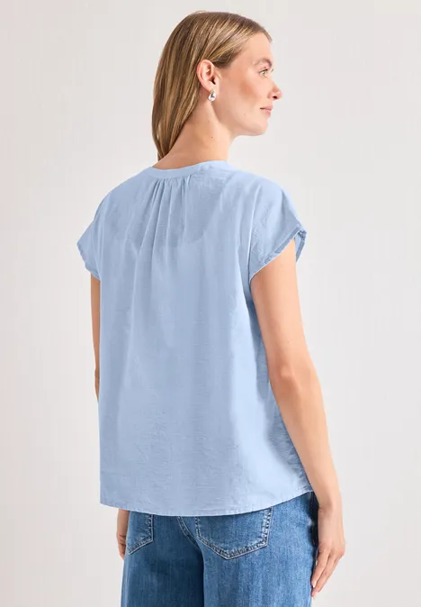 Vrouw van achteren in lichtblauw kort mouwshirt en spijkerbroek, licht zijwaarts gedraaid voor neutrale achtergrond.