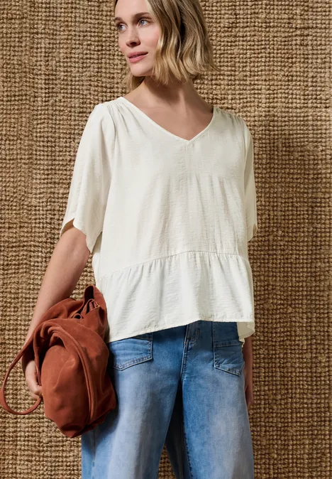 Kurzarm Bluse mit V-Neck und Schößchen off white Frau trägt weiße V-Ausschnitt-Bluse, blaue Jeans und roten Rucksack vor geflochtenem Hintergrund.