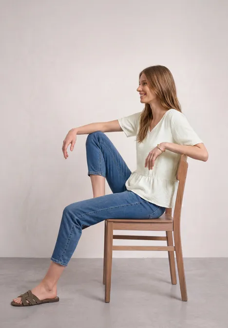 Kurzarm Bluse mit V-Neck und Schößchen off white Frau sitzt seitlich auf einem Holzstuhl, lehnt den Arm an die Rückenlehne, trägt Jeans und T-Shirt.