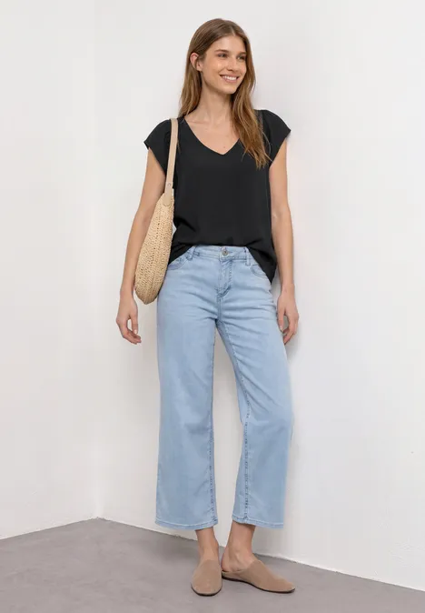 Dropped Shoulder Shirt mit V-Neck Black Frau steht in Zimmerecke an weißer Wand, trägt schwarzes V-Shirt, weite hellblaue Jeans, beige Schuhe und Strohtasche.