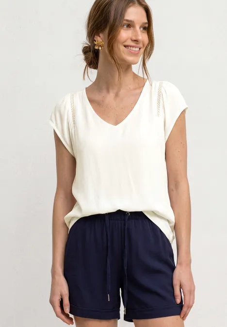 Frau in cremeweißem V-Neck-Shirt und dunkelblauen Shorts steht vor neutralem Hintergrund.