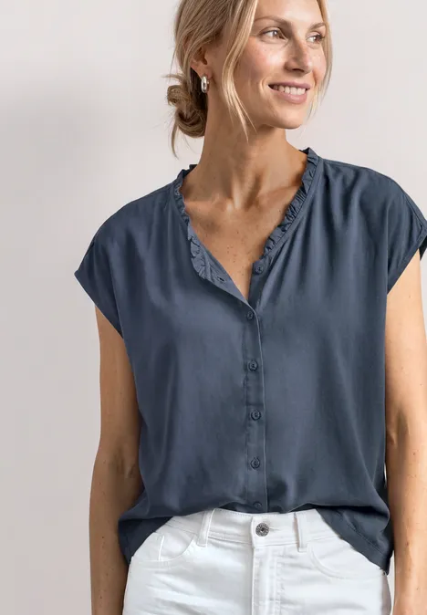 Vrouw draagt donkerblauwe, korte mouw blouse met ruches en een witte broek.