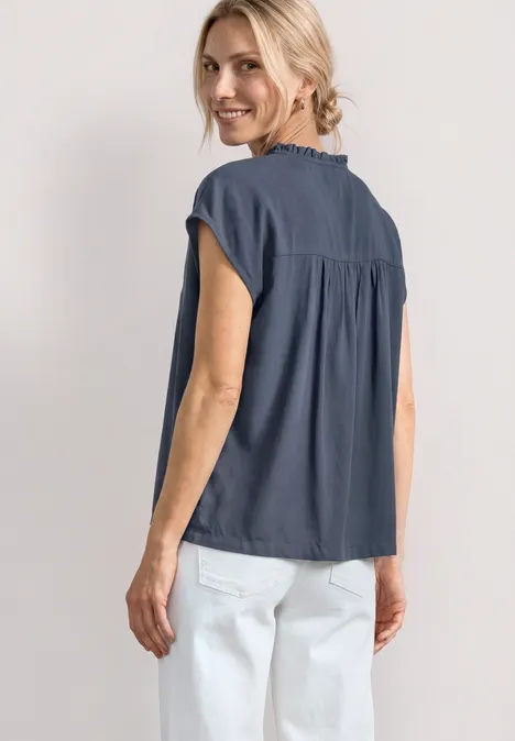 Vrouw van achteren, donkerblauw hemd met korte mouwen en witte broek, lichte achtergrond.