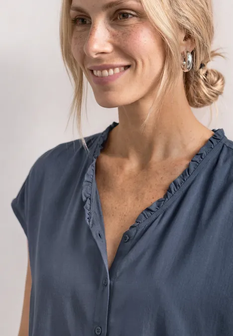 Blonde vrouw in een los blauw shirt met V-hals, glimlacht zachtjes opzij naar de camera.
