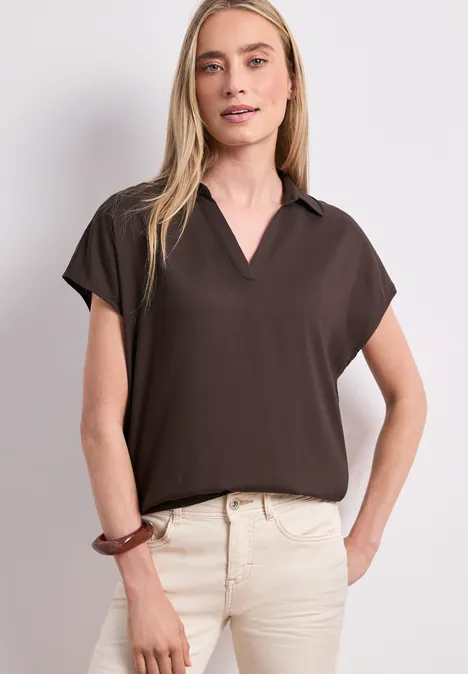 Frau trägt braunes Kurzarmshirt und beiges Hose, Hand an der Hüfte.