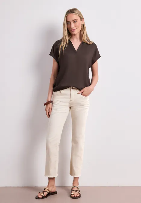 Eine Frau steht vor weißem Hintergrund, trägt braunes V-Shirt, helle Jeans und schwarze Sandalen.