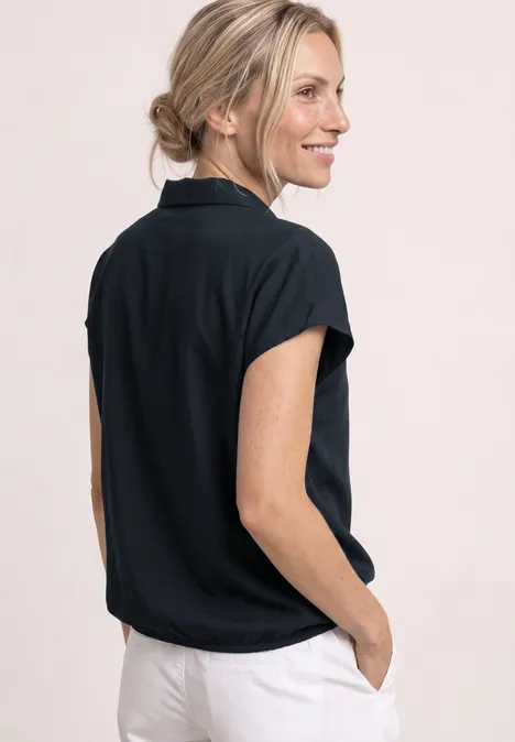 Achteraanzicht van een vrouw in een zwarte korte mouw blouse met kraag en lichte broek, handen in de zakken.