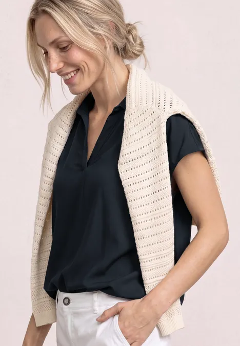 Vrouw in zwart blouse met crème gilet over de schouders, hand in de broekzak.
