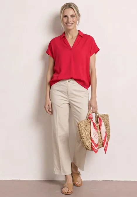 Frau in roter Bluse, weißer Hose und Basttasche mit Schal vor hellem Hintergrund.
