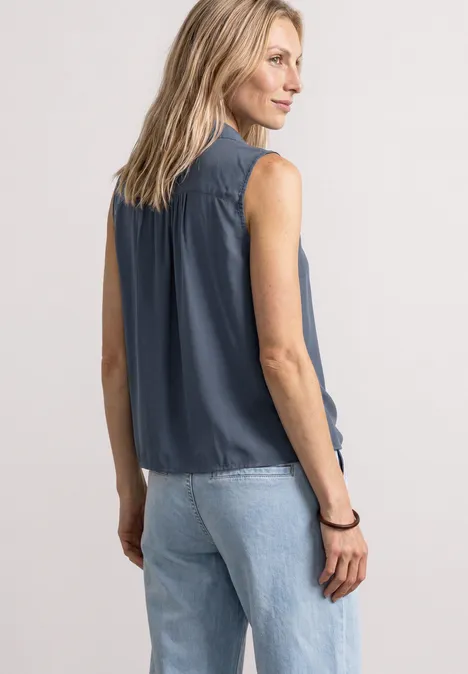 Blusentop mit Bowling-Kragen und Knoten ashen blue Frau in ärmelloser dunkelblauer Bluse und hellen Jeans von hinten fotografiert.