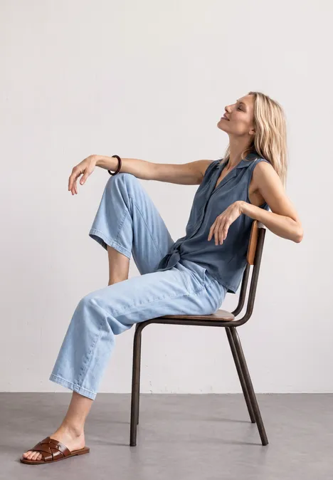 Blusentop mit Bowling-Kragen und Knoten ashen blue Frau sitzt entspannt seitlich auf Stuhl, trägt hellblaue Denim-Hose, mit erhobenem Blick, minimalistischer Hintergrund.