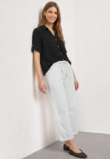 Vrouw leunt tegen witte muur, draagt zwarte blouse, witte wijde broek en zwarte ballerina’s.