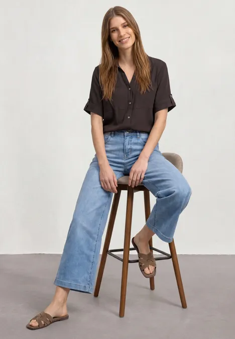 Kurzarm Hemdbluse mit Turn-Up-Details espresso brown Frau auf hohem Hocker, trägt dunkelbraunes Hemd, weite Jeans und Sandalen vor weißem Hintergrund.