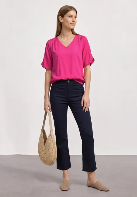 Frau in pinker Bluse und dunkelblauen Jeans mit Strohtasche und beigen Schuhen vor neutralem Hintergrund.