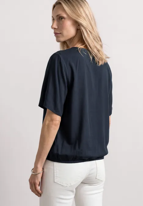 Blouseshirt met korte mouwen en V-hals shadowed navy Blondine gezien van achter, zwarte korte mouw top en witte spijkerbroek.