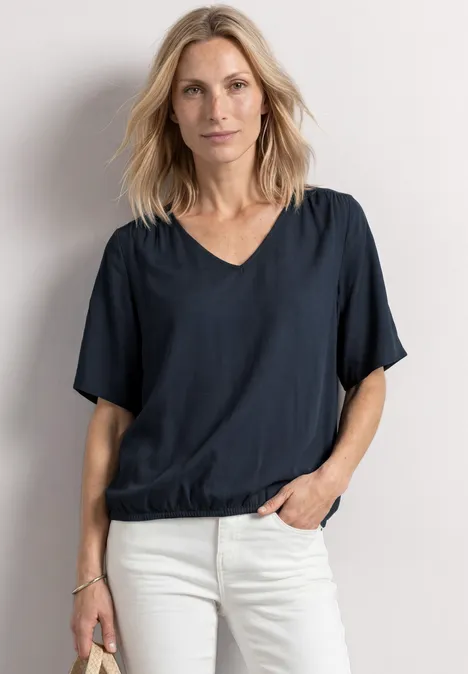 Blouseshirt met korte mouwen en V-hals shadowed navy Vrouw in donkerblauwe top en witte jeans, met één hand in de zak, staand tegen een neutrale achtergrond.