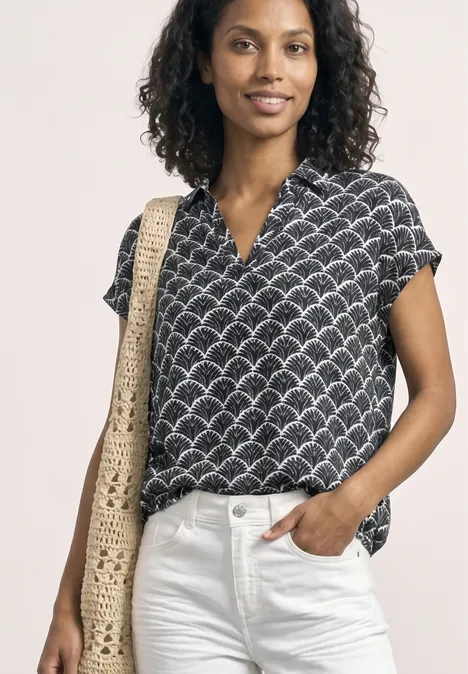 Eine Person trägt eine schwarze Bluse mit kurzem Arm und Muschel-Print, weiße Jeans und eine gehäkelte Strohtasche.