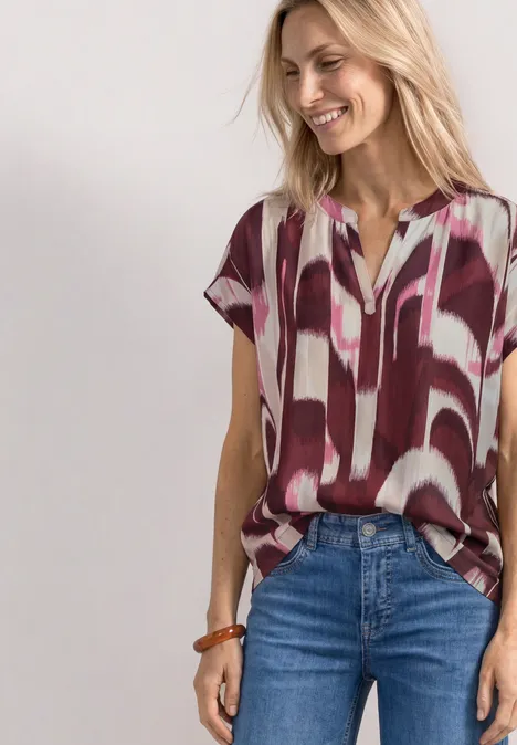 Vrouw draagt losvallende korte mouw blouse met abstract rood-wit dessin en jeans.