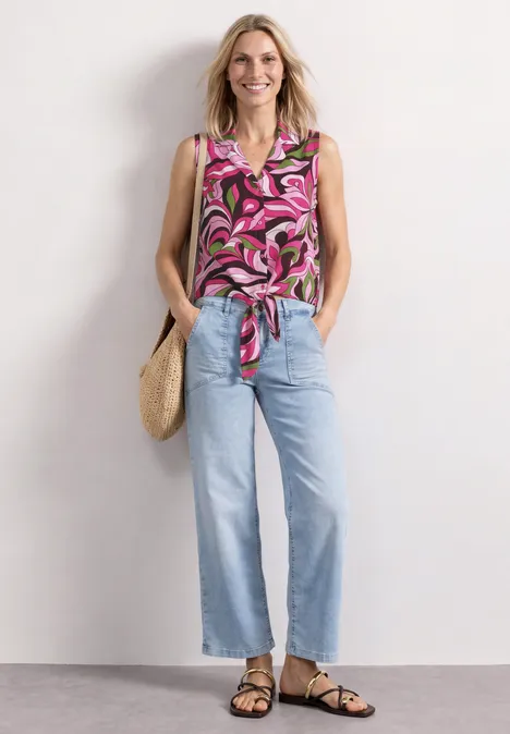 Blusentop mit Bowling-Kragen und Knoten magenta dream Frau in geblümter, pinker Bluse mit Knoten, blauen Jeans, Sandalen und Strohtasche vor weißem Hintergrund.