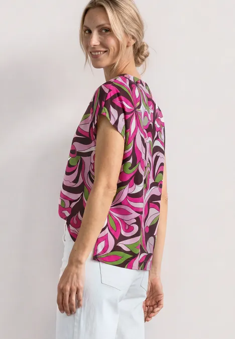 Vrouw in kleurrijke, korte mouwen top met abstracte swirlprint in roze, groen en zwart en witte broek.