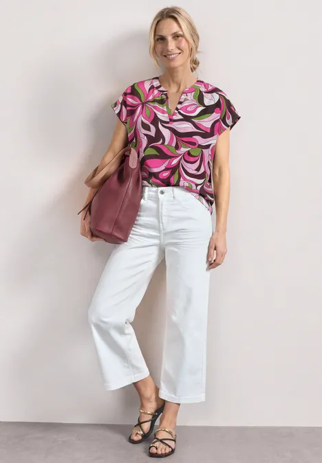 Vrouw met knot in roze dessinblouse, witte wijdere broek, zwarte sandalen en een grote roze tas.