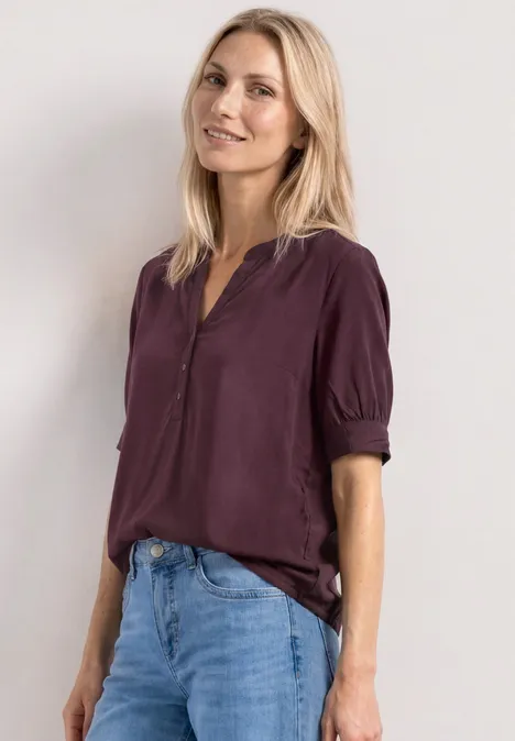 Een vrouw draagt een donkerpaarse blouse met opgerolde mouwen en een blauwe spijkerbroek.