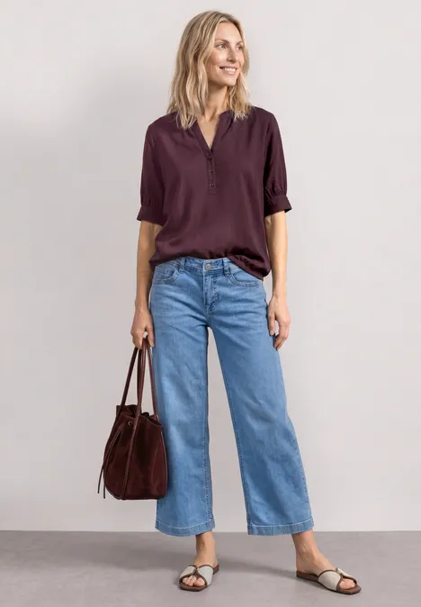 Vrouw draagt wijde lichtblauwe jeans, donkerpaarse blouse, zwarte sandalen en een bordeauxrode tas.