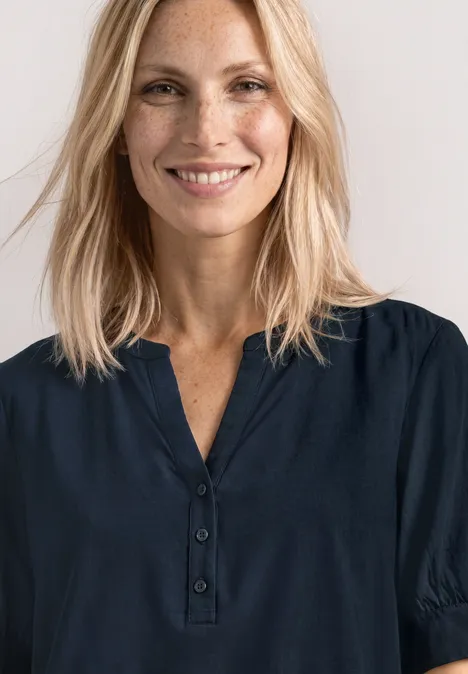 Een vrouw met halflang blond haar draagt een donkerblauw hemd met knopen, gezicht onscherp.