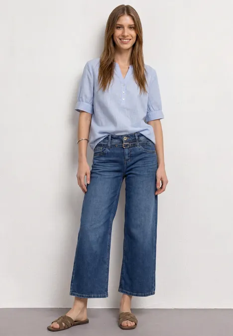 Eine lächelnde Frau in hellblauer Bluse, weiten Jeans und Sandalen vor weißem Hintergrund.