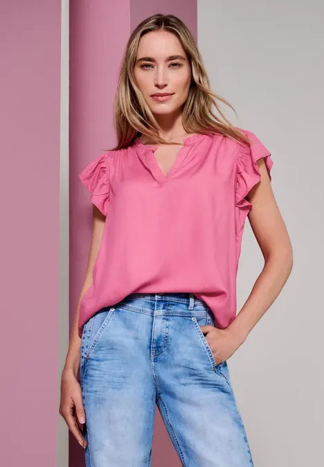 Frau in rosa Bluse und Jeans posiert vor zweifarbigem Hintergrund.