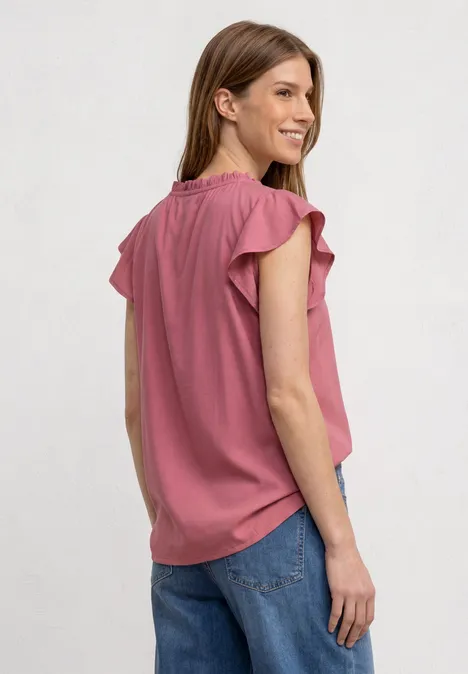 Frau trägt rosa Bluse mit Rüschenärmeln und blaue Jeans, seitliche Ansicht, lächelnd.