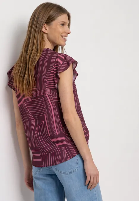 Eine Frau von hinten in blau-pink gemusterter Bluse mit Rüschenärmeln und Jeans, lächelnd zur Seite blickend.