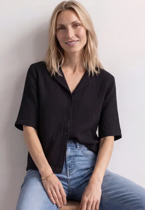 Musselin Bluse mit Bowling-Kragen Black Eine lächelnde Frau mit blonder Mähne, trägt schwarzes Hemd und Jeans, sitzt vor weißer Wand.