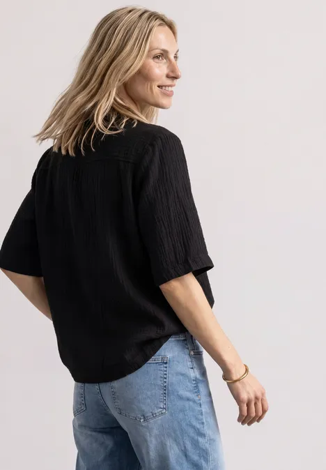 Musselin Bluse mit Bowling-Kragen Black Blonde Frau im Halbprofil mit schwarzem, lockerem Oberteil und blauer Jeans vor hellem Hintergrund.