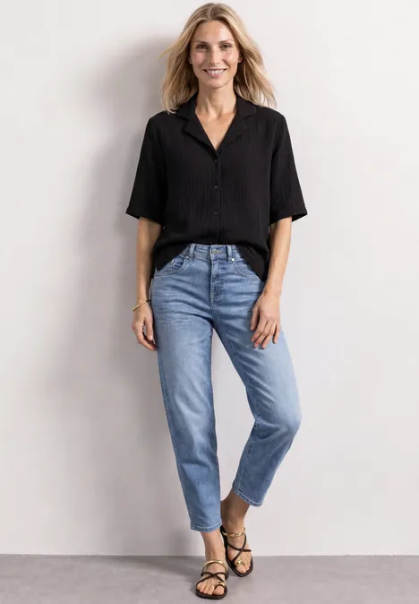 Musselin Bluse mit Bowling-Kragen Black Frau in schwarzer Bluse, Jeans und Sandalen vor neutralem Hintergrund.