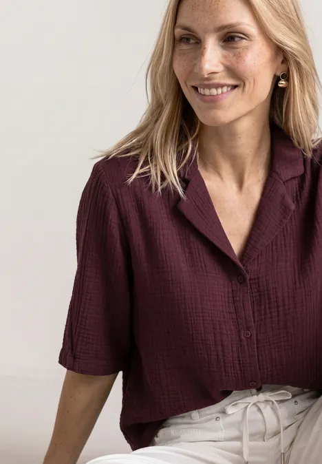 Frau mit blondem Haar, lächelnd, trägt eine weinrote Bluse mit Taschen und halblangen Ärmeln.