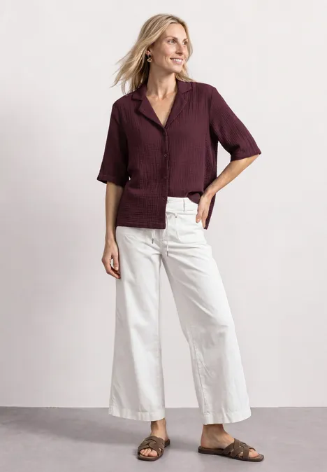 Frau in bordeauxfarbener Bluse, weißen Culotte-Hosen und braunen Sandalen, lächelnd vor neutralem Hintergrund.