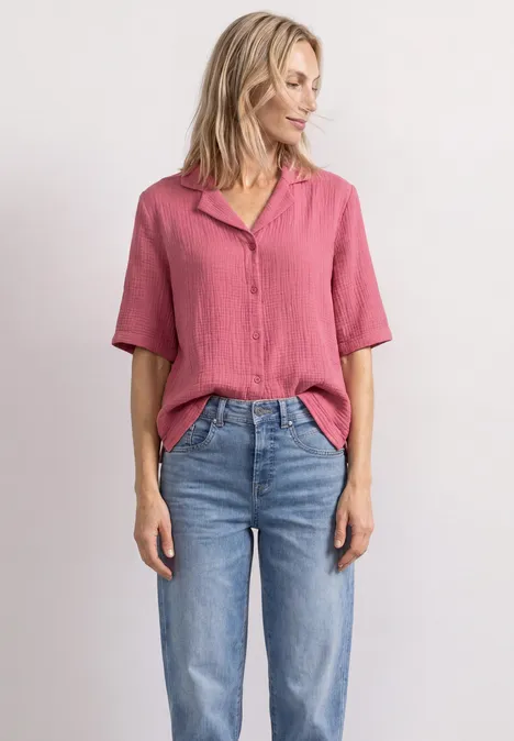 Frau in rosa Bluse und blauen Jeans steht lässig vor weißem Hintergrund.