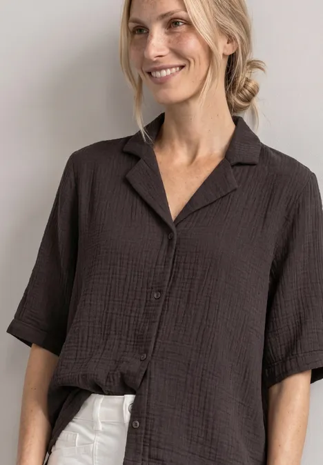 Mousselineblouse met bowlingkraag black coffee Vrouw glimlachend in een losse, zwarte gestructureerde blouse met korte mouwen en een witte broek.