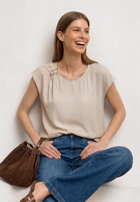 Blusenshirt mit Rundhals und Schnallendetail clay sand Frau sitzt vor heller Wand, trägt beiges Top, blaue Jeans und braune Tasche.