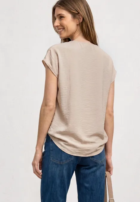 Blusenshirt mit Rundhals und Schnallendetail clay sand Eine Frau steht von hinten in beigem T-Shirt, blauen Jeans und hält eine Tasche.