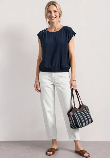 Blusenshirt mit Rundhals und Schnallendetail darkest blue Frau in marineblauer Bluse, weißen Hosen, flachen Sandalen und gestreifter Tasche vor hellem Hintergrund.