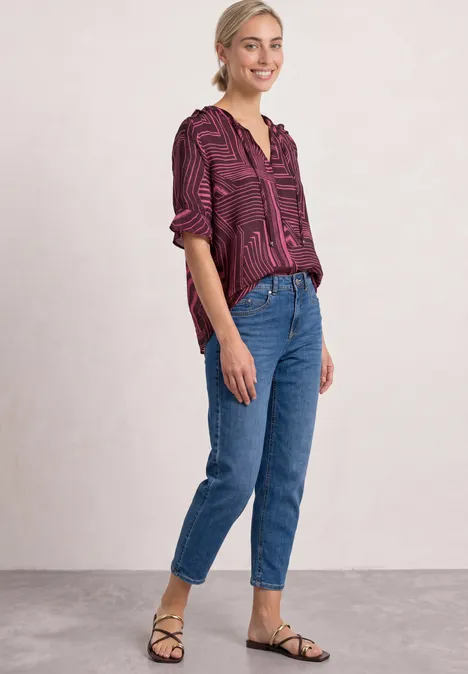 Blouse met korte mouwen, split in de hals en print blush berry Vrouw in bordeauxblouse en spijkerbroek, staand voor neutrale achtergrond, ontspannen glimlach.
