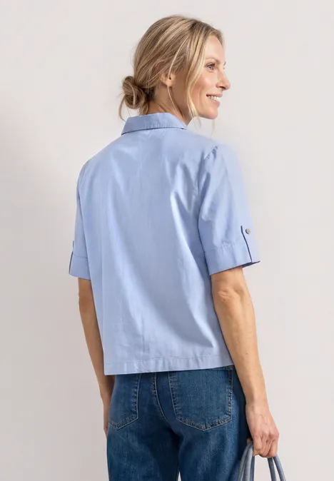 Rückansicht einer Frau in hellblauer, kurzärmliger Bluse und blauen Jeans vor weißem Hintergrund.