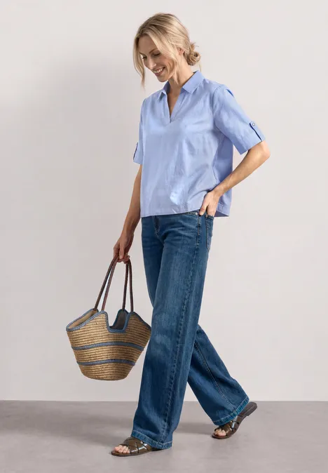 Frau in hellblauem Kurzarmhemd, weiten Jeans und Basttasche, entspannt im Gehen.