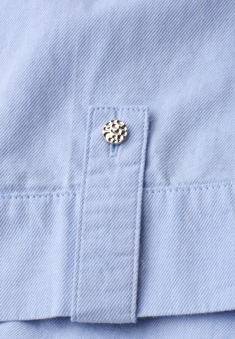 Detail eines hellblauen Hemdplackets mit einem verzierten Metall-Druckknopf.