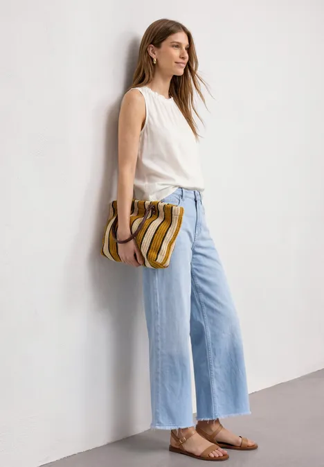 Frau in weißem Top, hellblauen weiten Jeans und braunen Sandalen lehnt an weißer Wand und hält gestreifte Clutch.