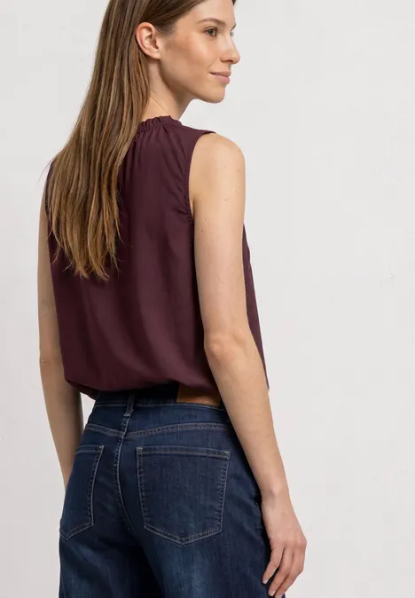 Eine Frau von hinten in einer ärmellosen bordeauxfarbenen Bluse und blauen Jeans.